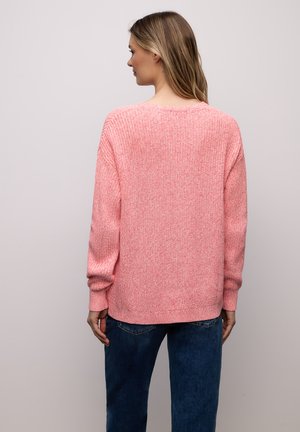Femme de dos portant un pull en maille rose et un jean bleu sur un fond gris clair uni.