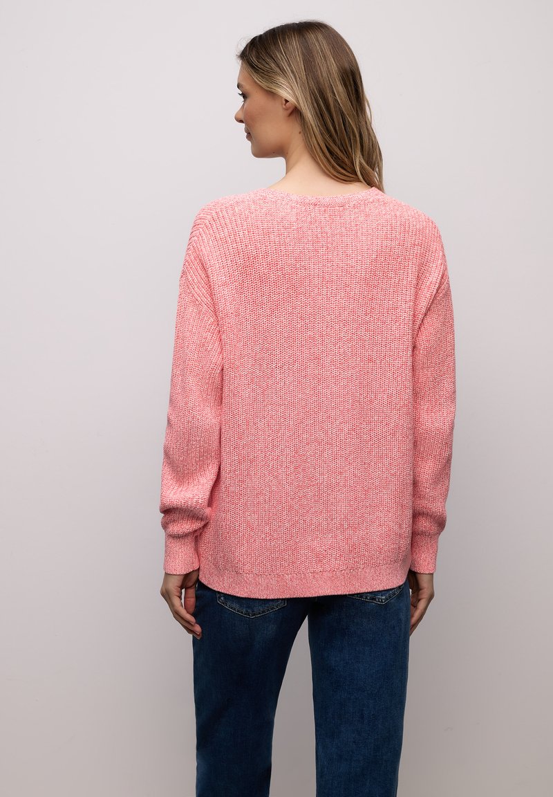 Femme de dos portant un pull en maille rose et un jean bleu sur un fond gris clair uni.