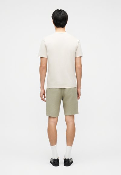 Άνδρας που στέκεται με την πλάτη γυρισμένη, φοράει απλό μπεζ μπλουζάκι, ανοιχτόχρωμο ελιάς σορτς, λευκές κάλτσες και μαύρα παπούτσια σε λευκό φόντο.