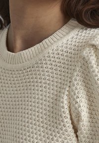 Cremfarbener Strickpullover mit einem strukturierten, offenen Strickmuster und einem gerippten Ausschnitt. Der Stoff hat ein weiches, natürliches Aussehen.