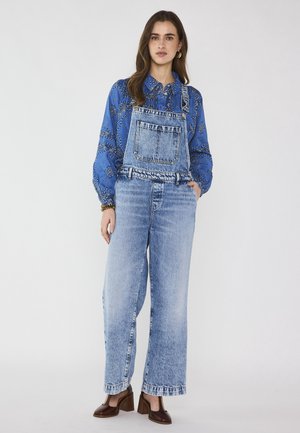 Donna che indossa una camicetta blu con disegni e una salopette di jeans azzurro chiaro, in piedi con una mano in tasca e scarpe con tacco a blocco marrone.