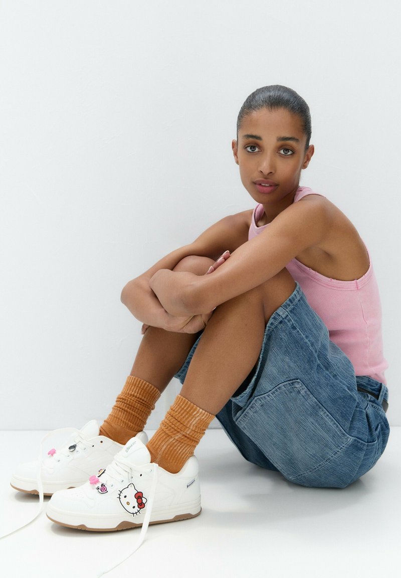 Baskets blanches avec des motifs Hello Kitty, semelles beige, accompagnées de chaussettes côtelées orange. Shorts en denim et débardeur rose clair, assis sur un fond neutre.