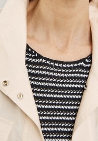 Beżowa, lekka kurtka z zatrzaskami, nałożona na czarno-biały prążkowany sweterek z fakturowanym wzorem i falistymi krawędziami.