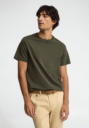 Uomo che indossa una t-shirt verde oliva a maniche corte e pantaloni beige con cintura marrone, che guarda leggermente verso sinistra su uno sfondo semplice.