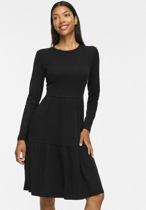 Femme aux longs cheveux foncés portant une robe noire à manches longues longueur genou, souriante et posant devant un fond blanc uni.