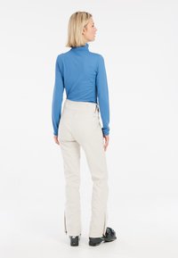 Maglia a maniche lunghe di colore azzurro chiaro abbinata a pantaloni da sci color crema con zip alle caviglie e scarponi da sci neri. Design liscio e aderente.