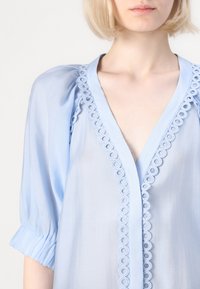 Lichtblauw shirt met pofmouwen, een diepe V-hals en decoratieve kantafwerking met cirkelpatronen aan de voorkant. Zachte, doorschijnende stof.