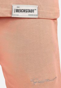 Tessuto color pesca chiaro con una texture liscia. L'etichetta mostra il nome del marchio "REICHSTADT" e una firma ricamata in argento.