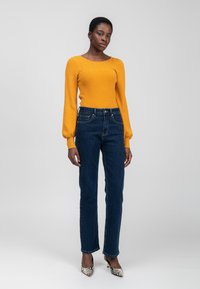 Eine Frau steht in einem senfgelben Strickpullover, dunkblauen, geraden Jeans und hochhackigen Schuhen mit Schlangenledermuster vor einem schlichten grauen Hintergrund.