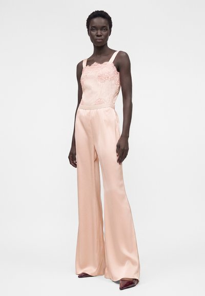 Γυναίκα που φοράει ένα ανοιχτό ροζ δαντελένιο τοπ που είναι μπαγκάκια μέσα σε αντίστοιχο φαρδύ μακρύ σατέν παντελόνι, συνδυασμένο με μυτερές σκούρες μπορντό παπούτσια.