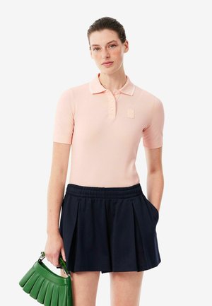 Vrouw draagt een lichtroze poloshirt met korte mouwen en een marineblauwe geplooide rok, houdt een groene geplooide handtas in haar linkerhand.
