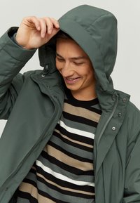Gray puffer jacket with a hood, featuring a zip closure and buttons, worn over a striped sweater in black, beige, and gray colors.