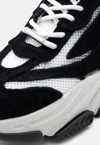 Κοντινή λήψη μαύρου και λευκού αθλητικού παπουτσιού με πάνελ από δίχτυ, επικαλύψεις σουέντ, λευκά κορδόνια και ασημένια ακροφύσια με ανεξίτηλο το όνομα "Steve Madden".