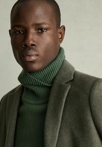 Pulover din tricot verde cu guler înalt, purtat sub un sacou din lână gri. Puloverul are o textură cu riburi, în timp ce sacoul are o finisare netedă.