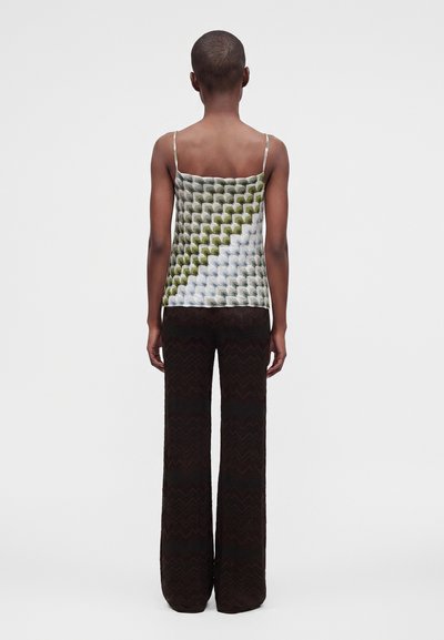 Personne aux cheveux courts portant un haut sans manches à motif et un pantalon large foncé, debout de dos sur un fond uni.