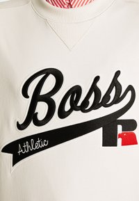 Sweatshirt blanc avec un col rond côtelé, arborant un logo "Boss" brodé en noir et rouge ainsi qu'une bannière "Athletic" en couture blanche.
