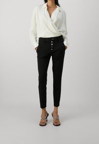 Weiße, langärmelige Wickelbluse kombiniert mit schwarzen, maßgeschneiderten Hosen mit Knopfleiste. Dazu schwarze High-Heel-Sandalen als Accessoires.