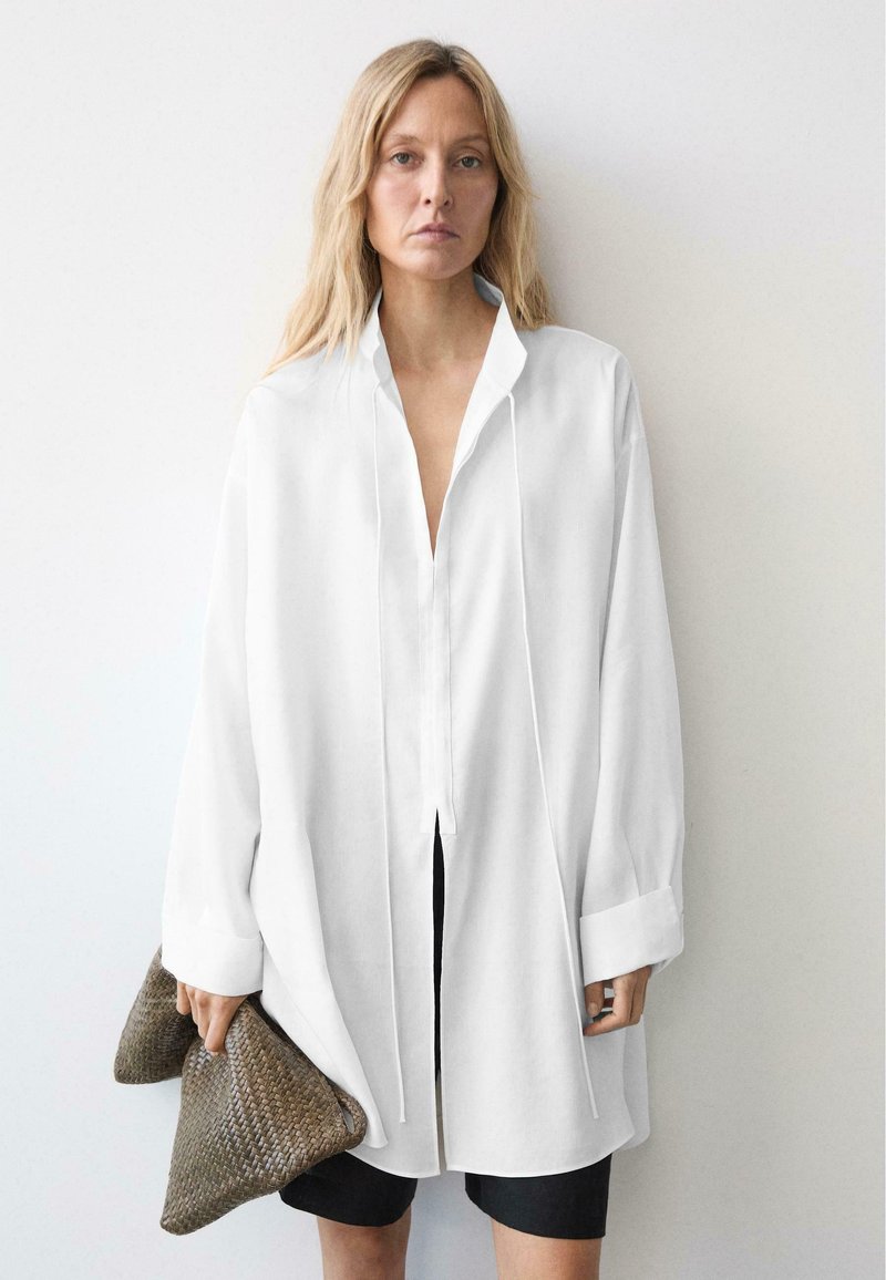 Kvinde med langt blondt hår iført en stor hvid skjorte og sorte shorts, holder en stor tekstureret brun clutch op ad en ensfarvet væg.