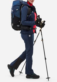 Veste de plein air marine avec une doublure intérieure rouge, pantalon assorti en marine, gants noirs et sac à dos Millet. Chaussures de randonnée noires avec des accents bleu-vert.