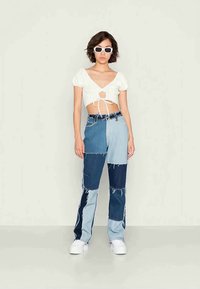 Top branco curto com mangas bufantes e amarração ao centro, combinado com calças de ganga em patchwork multicolorido. Modelo usa óculos de sol brancos e ténis.