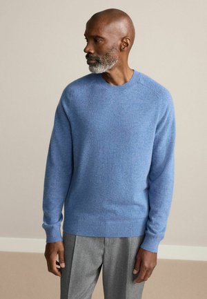 Helles blaues Rundhals-Pullover aus weichem Strickstoff, mit langen Ärmeln und gerippten Bündchen. Kombiniert mit grauen maßgeschneiderten Hosen.