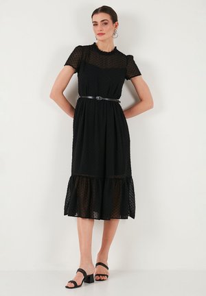 Black dress with sheer dotted fabric, short sleeves, ruffled hem, cinched waist with a thin belt, complemented by square-heeled sandals.