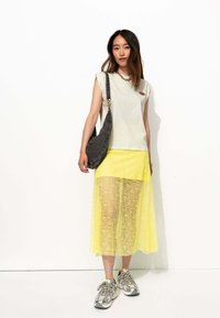 Sac d'épaule gris, haut sans manches gris clair avec logo, jupe maxi en dentelle jaune, et baskets grises avec accents réfléchissants et lacets à motifs.