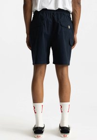 Marineblauwe cargoshorts met een elastische taille, zijzakken en een labeldetail. Gecombineerd met witte sokken met rode letters en zwarte schoenen.