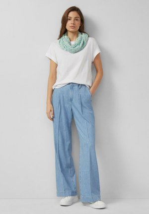 Femme debout contre un mur gris, portant un t-shirt blanc, un jean large bleu clair, des baskets blanches et une écharpe infinie bleue à motifs.