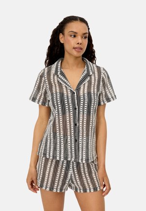 Mujer vestida con una camisa de manga corta abotonada con un patrón en negro, blanco y marrón, y shorts a juego, de pie y mirando hacia la derecha.