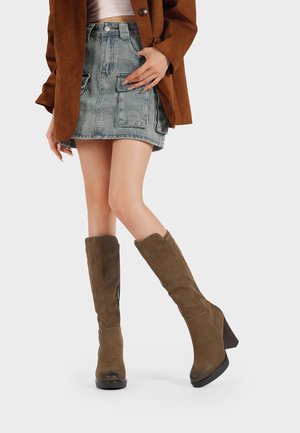 Donna che indossa stivali marroni in camoscio al ginocchio, una gonna di jeans blu con grandi tasche, una giacca marrone e una maglietta bianca su sfondo bianco.