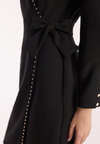 Czarna sukienka z bocznym detalem w postaci kokardy, ozdobiona złotymi guzikami i linią złotych ćwieków biegnącą wzdłuż szwu. Gładka tekstura.