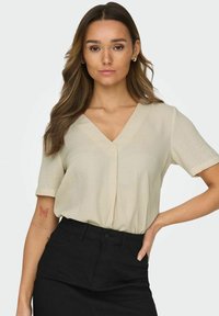 Beige blouse met korte mouwen en een V-hals, met een losse pasvorm en lichte textuur. Gecombineerd met een zwarte hoog-getailleerde rok.