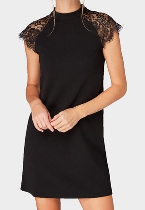 Vrouw draagt een zwarte mouwloze jurk met kanten kapmouwen, staat met handen losjes in elkaar gevouwen ter hoogte van de taille.