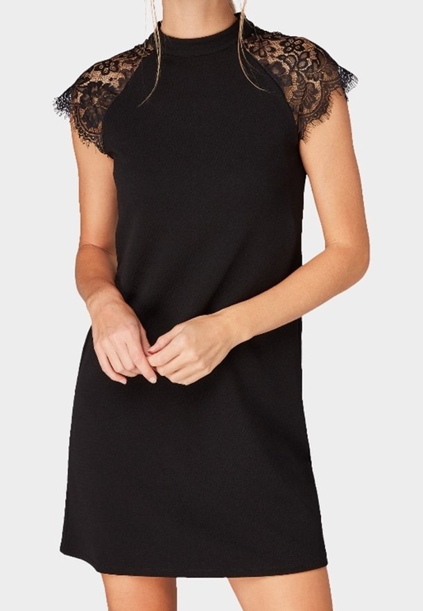 Femme portant une robe noire sans manches avec des manches en dentelle couvrant les épaules, debout les mains légèrement croisées à la taille.