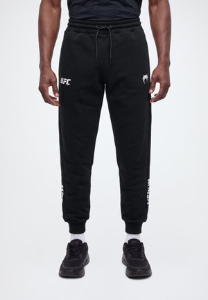 El modelo lleva unos pantalones de chándal negros de UFC con cintura con cordón y logotipos blancos, combinados con zapatillas deportivas negras y calcetines blancos sobre un fondo liso.