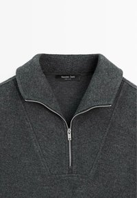 Tmavě šedý fleecový pulovr s límcem na zip a výstřihem ve tvaru písmene V. Má hladkou texturu a jemné šicí detaily.