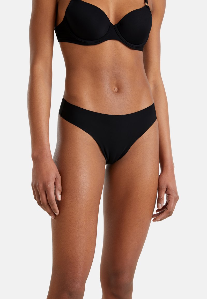 Femme portant un soutien-gorge noir à armatures et un bikini noir sans coutures, se tenant devant un fond clair.