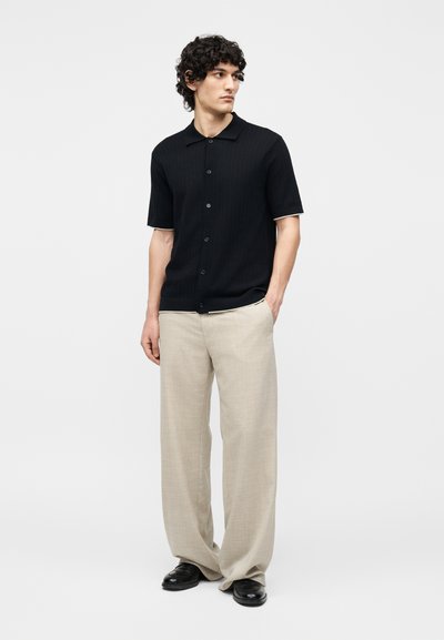 Νέος άνδρας που φοράει μαύρο πουκάμισο με κοντά μανίκια και κουμπιά, μπεζ παντελόνι με φαρδιά μπατζάκια και μαύρα παπούτσια, στέκεται με το ένα χέρι στην τσέπη.