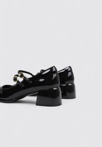 Zapatos tipo Mary Jane de charol negro con punta redondeada, pequeño tacón cuadrado y dos hebillas decorativas en tono dorado en la correa.