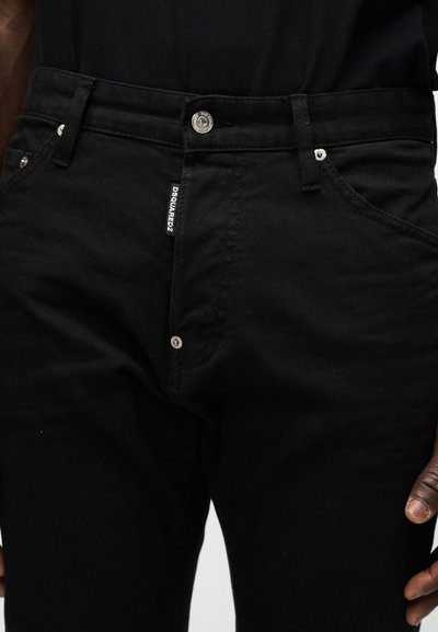 Κοντινό πλάνο ατόμου που φοράει μαύρα τζιν με μεταλλικό κουμπί και πριτσίνια, δείχνοντας την ετικέτα της μάρκας Dsquared2 στο φερμουάρ.