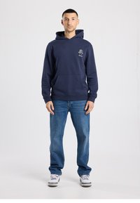 Navy blue hoodie with a front pocket and a drawstring hood, featuring a small tree logo on the left chest. Worn with blue jeans and sneakers.