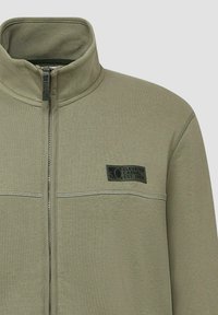 Oliwkowa, rozpinana bluza z wysokim kołnierzem. Posiada płaskie szwy, materiałową naszywkę z logo oraz gładkie, teksturowane wykończenie.