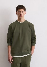 Sweatshirt vert olive avec des manches longues, un col rond et une texture douce, superposé à une chemise blanche et associé à un pantalon assorti.