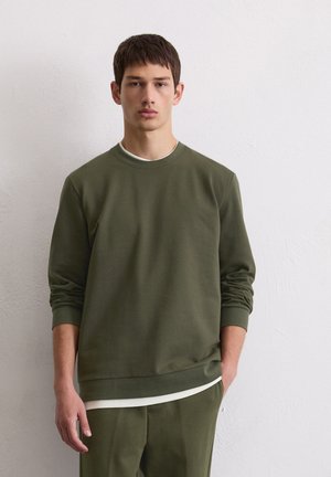 Olijfgroen sweatshirt met lange mouwen, een ronde halslijn en een zachte textuur, in laagjes over een wit shirt en gecombineerd met bijpassende broek.