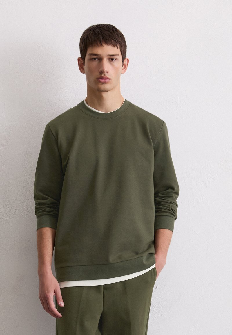 Sweatshirt vert olive avec des manches longues, un col rond et une texture douce, superposé à une chemise blanche et associé à un pantalon assorti.