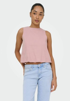 Femme portant un haut court rose sans manches et un jean taille haute bleu clair, debout avec une main derrière le dos.