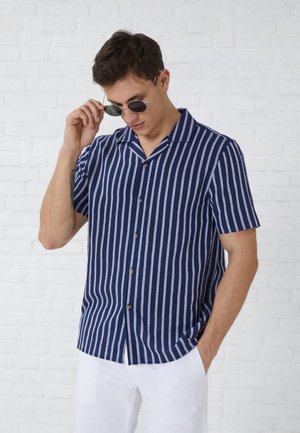 Άνδρας με κοντομάνικο πουκάμισο με κατακόρυφες μπλε σκούρες και λευκές ρίγες και λευκό παντελόνι, προσαρμόζει στρογγυλά μαύρα γυαλιά ηλίου μπροστά από λευκό τούβλινο τοίχο.