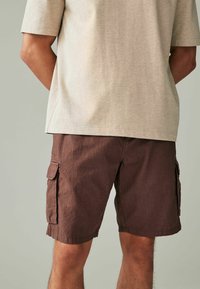 Braune Cargo-Shorts mit seitlichen Taschen, aus strukturiertem Stoff. Kombiniert mit einem hellgrauen T-Shirt mit kurzen Ärmeln, von hinten betrachtet.