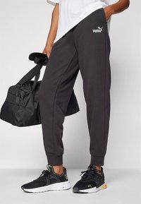 Czarne zwężane spodnie dresowe z bocznymi szwami, z małym białym logo Puma. W połączeniu z czarnymi butami sportowymi i czarną torbą sportową.